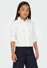 Camicia bianca in cotone con maniche arrotolate, caratterizzata da piccoli elementi decorativi e un design accorciato. Abbinata a pantaloni blu navy.