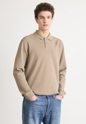 Beige long-sleeve polo shirt with a textured pattern, two-button collar, and subtle logo on the chest. Paired with blue denim jeans.