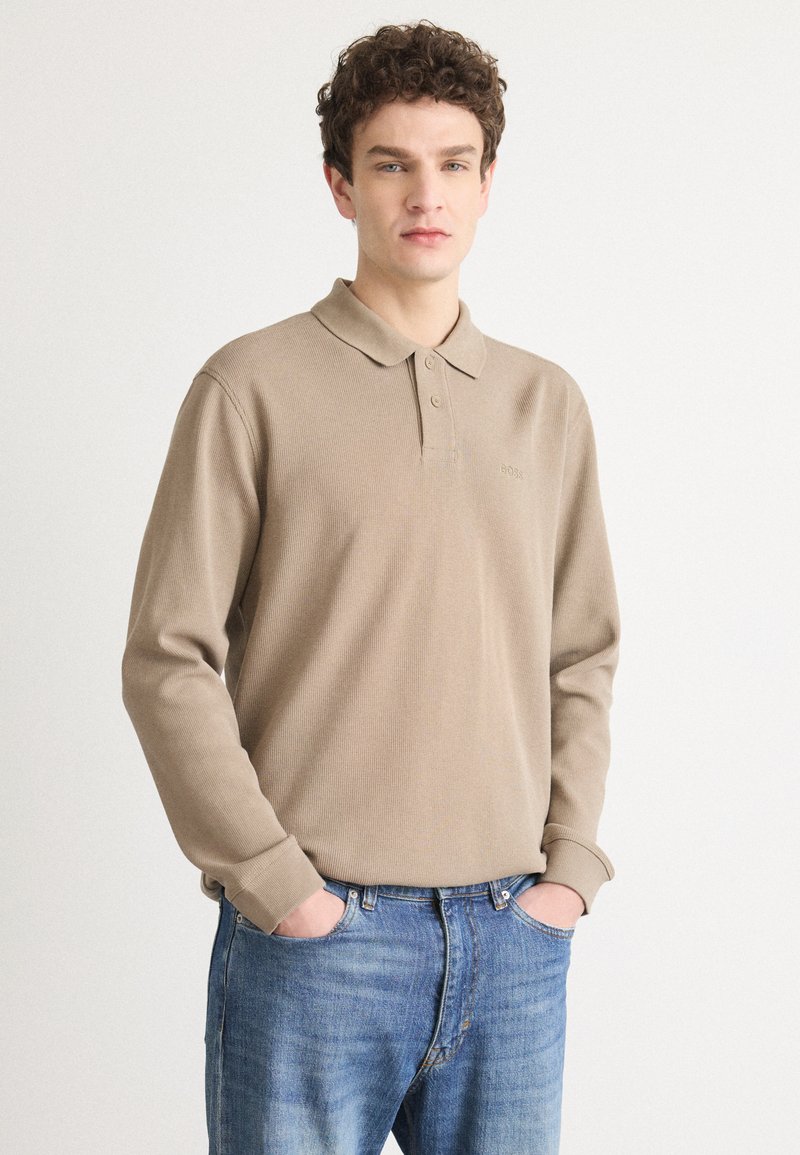 Beige langermet polo skjorte med et teksturert mønster, krage med to knapper og diskret logo på brystet. Kombinert med blå denimjeans.