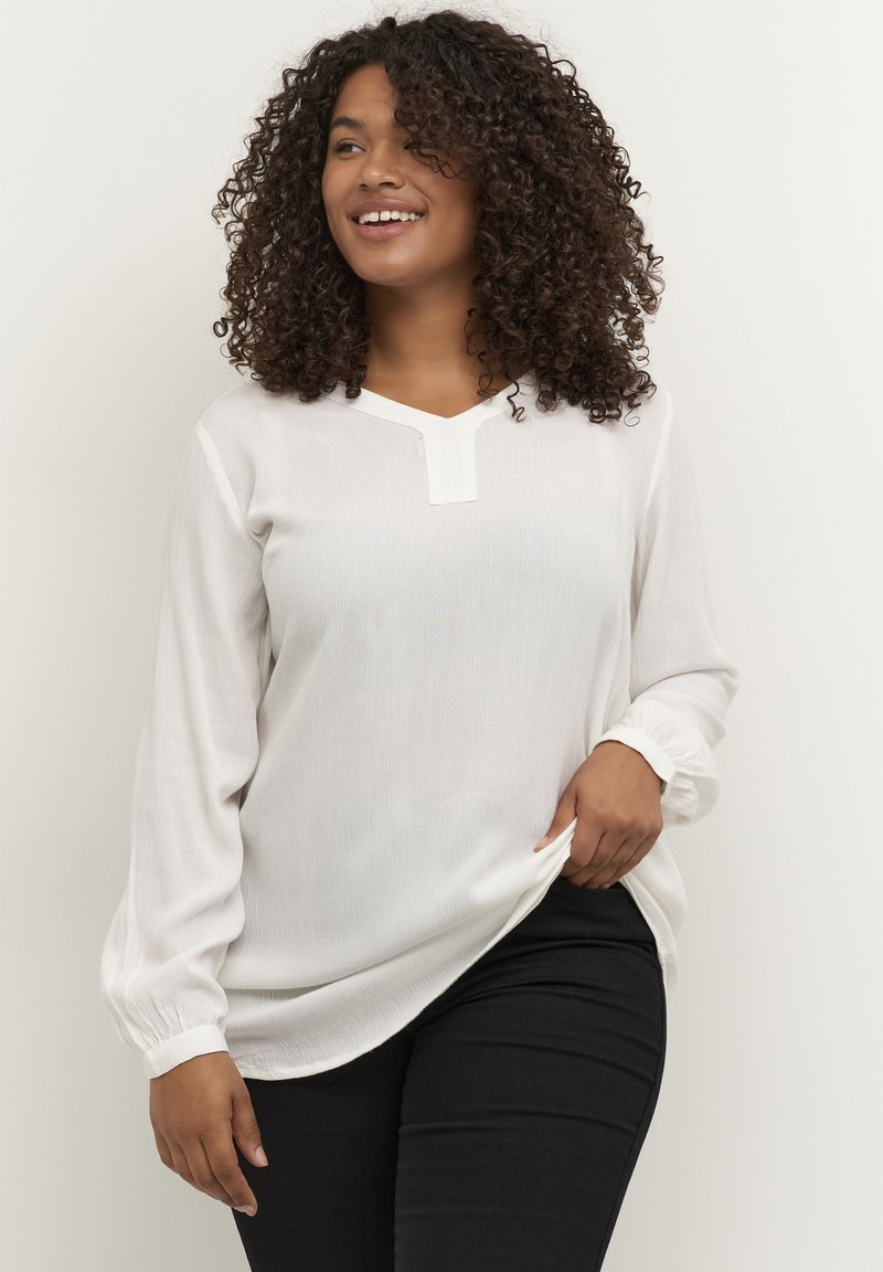 Weiße Bluse mit langen Ärmeln, V-Ausschnitt und strukturiertem Stoff. Mit elastischen Bündchen und lockerer Passform. Kombiniert mit schwarzen Slim-Fit-Hosen.