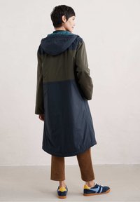 Waterdichte jas in groen en marineblauw; heeft een capuchon, lange mouwen en een middellange uitstraling. Gecombineerd met een paar bruine corduroy broeken en sneakers.