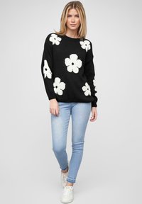 Pull en tricot noir avec un grand motif floral blanc, col rond et manches longues, associé à un jean skinny bleu clair et des baskets blanches.