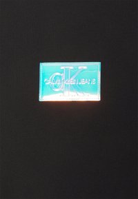 Niebieska prostokątna naszywka z wytłoczonymi na czarnym materiale literami "CK" i napisem "Calvin Klein Jeans".