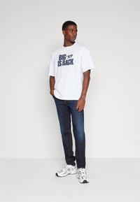 Hombre de pie con las manos en los bolsillos, vistiendo una camiseta blanca con el texto "BIG IS BACK", jeans azul oscuro y zapatillas blancas, contra un fondo liso.