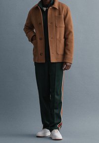 Braune Jacke aus Wollmischung mit zwei vorderen Taschen, darunter dunkelgrüner Trainingsanzug, grüne Hose mit weißen und orangefarbenen Streifen, weiße Schuhe.