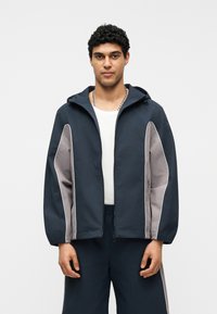 Άνδρας που φοράει μπλε ναυτικό και γκρι φούτερ με κουκούλα και φερμουάρ πάνω από λευκό πουκάμισο και ταιριαστό μπλε ναυτικό παντελόνι, στέκεται μπροστά σε απλό λευκό φόντο.