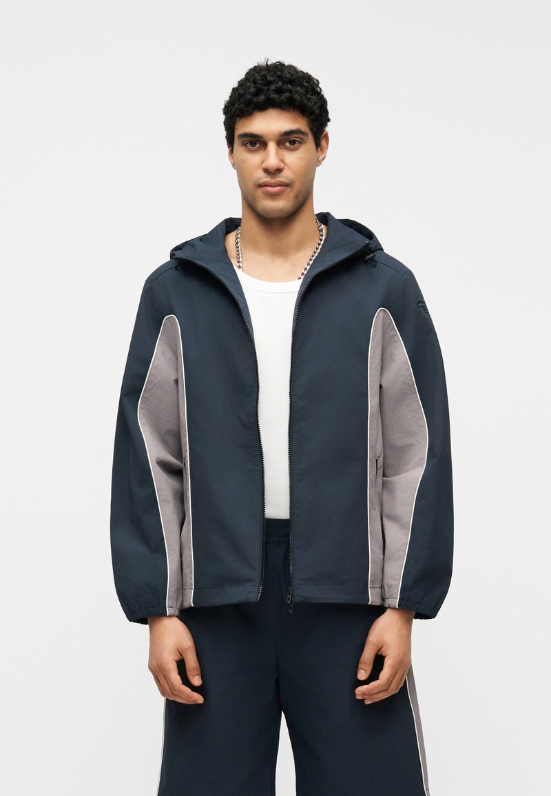 Άνδρας που φοράει μπλε ναυτικό και γκρι φούτερ με κουκούλα και φερμουάρ πάνω από λευκό πουκάμισο και ταιριαστό μπλε ναυτικό παντελόνι, στέκεται μπροστά σε απλό λευκό φόντο.