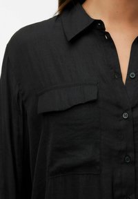 Camicia nera a maniche lunghe con bottoni, colletto, tasca a patta sul petto e cuciture visibili sulla parte frontale e sulle spalle.