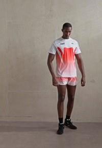 Man wearing white and orange athletic shirt and shorts, standing on gray floor against a plain concrete wall.