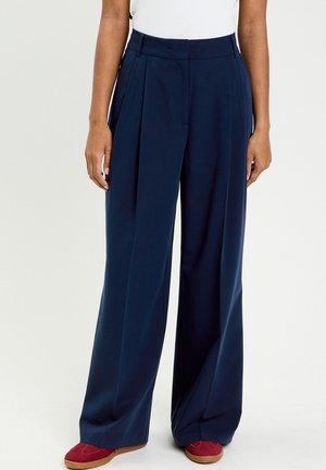 Persoon die marineblauwe wijd-uitlopende broek met plooien en rode suède schoenen draagt, staand tegen een effen achtergrond.