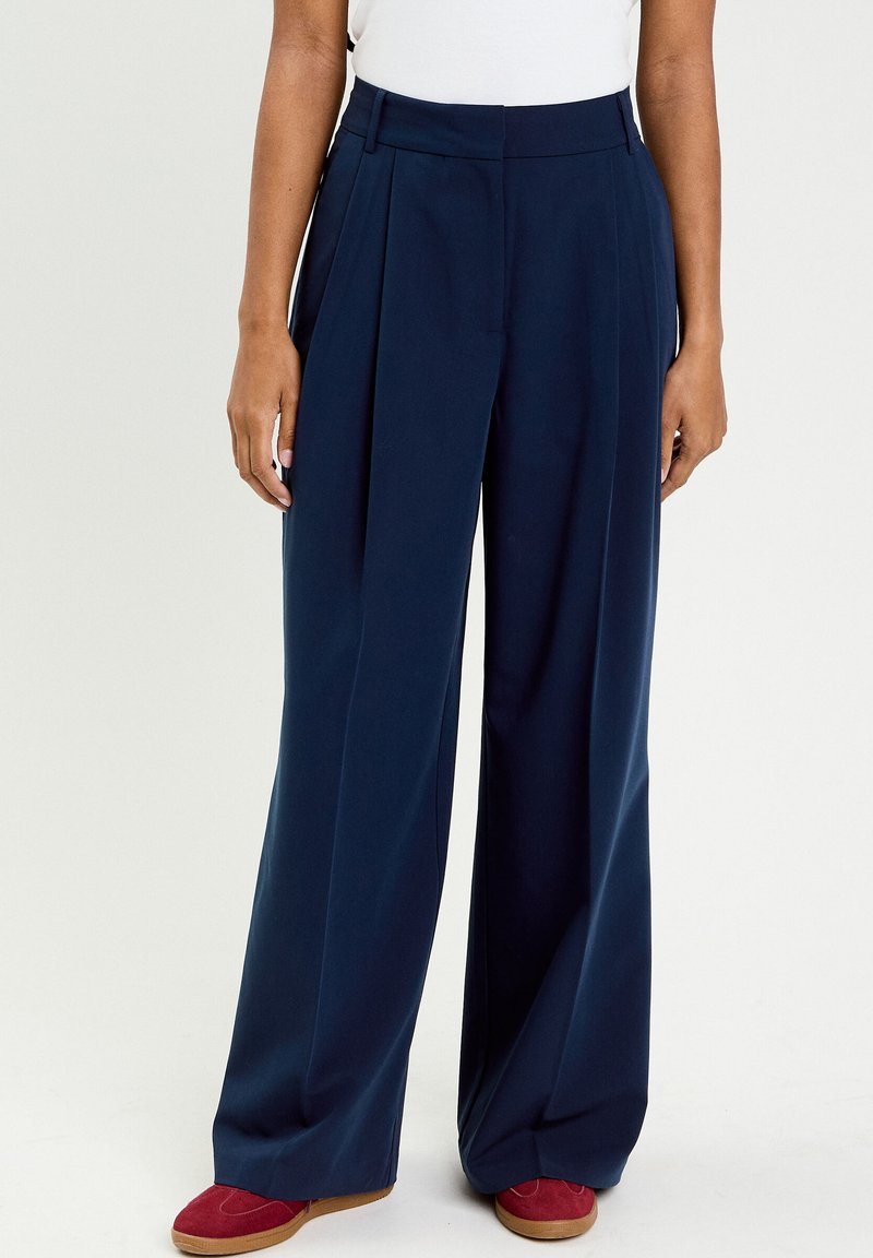 Persona che indossa pantaloni a gamba larga blu navy con pieghe e scarpe rosse in camoscio, in piedi contro uno sfondo uniforme.