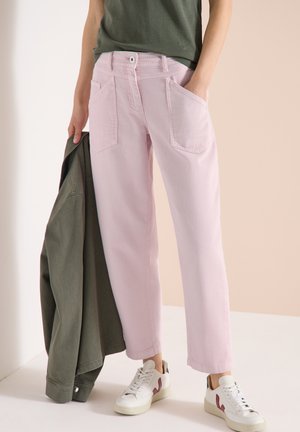 Persoon gekleed in lichtroze broek met wijde pijpen en witte sneakers, die een groene jas in één hand vasthoudt, staand tegen een neutrale achtergrond.