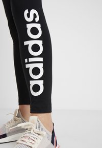 Czarne legginsy z dużym białym logo "adidas" po boku. W połączeniu z wielokolorowym obuwiem sportowym; widoczny oddychający materiał.