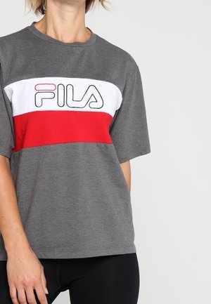 Person trägt ein graues FILA T-Shirt mit weißen und roten horizontalen Streifen und schwarze Hose vor einem schlichten hellen Hintergrund.