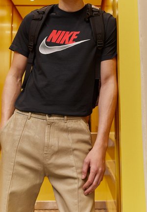 Personne portant un t-shirt Nike noir, un pantalon beige et des bretelles de sac à dos noires, debout contre un mur jaune à l'intérieur.
