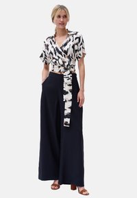 Koszula z krótkim rękawem w kolorze kremowym z czarnym i taupe abstrakcyjnym wzorem, zestawiona z wysokim stanem granatowymi spodniami w szerokim kroju oraz brązowymi sandałami na obcasie.