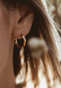 Goldene Creole mit einem verdrehten Design und einer reflektierenden Oberfläche. Vor einem verschwommenen Hintergrund mit sanfter, natürlicher Beleuchtung.