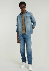 Camicia di jeans azzurro chiaro, parzialmente abbottonata sopra una maglietta polo beige. Indossata con jeans azzurro chiaro con dettagli in patch e scarpe bianche.