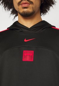 Homme portant un sweat à capuche noir avec le logo Nike rouge et le logo rouge des Chicago Bulls sur la poitrine, avec vue partielle du visage et du cou.