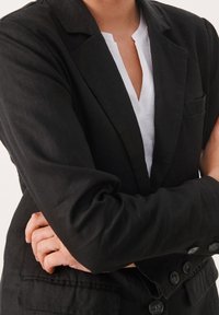 Schwarzer Leinenblazer mit Reverskragen, einer einzelnen Brusttasche und geknöpften Manschetten. Getragen über einem weißen Hemd mit V-Ausschnitt.