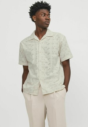 Camisa de manga corta en crema claro, con intrincados patrones cortados y un ajuste relajado, complementada con un cuello clásico y botones en la parte delantera.