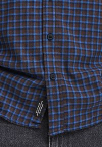 Bruin en blauw geruite button-up shirt met een zachte textuur, voorzien van zwarte knopen en een label dat aan de zoom is weergegeven.