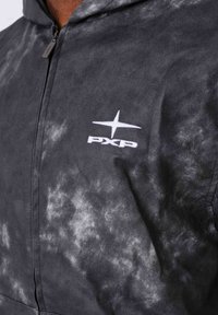 Primo piano di una felpa con zip in tie-dye nero e grigio con logo PXP ricamato in bianco e stella sul petto.