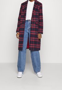 Henkilö, joka on pukeutunut punaisesta ja laivastonsinisestä ruudullisesta pitkistä takista valkoisen nappipaidan, löysien sinisten farkkujen ja vaaleansinisin yksityiskohdin varustettujen valkoisten lenkkareiden päälle.