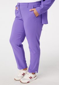 Paarse op maat gemaakte broek met zijzakken, taps toelopende pasvorm, elastische tailleband en eenvoudige zoom. Gedragen met witte sneakers met bordeauxrode accenten.