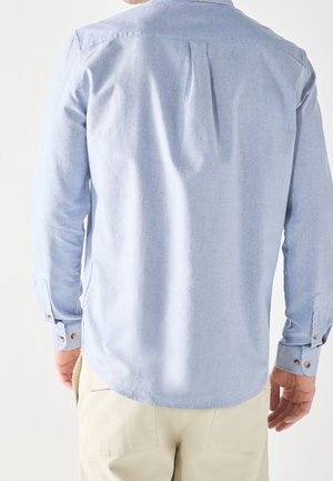 Homme portant une chemise bleu clair à manches longues et un pantalon beige, vu de dos sur un fond blanc uni.