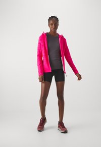 Bright pink zip-up hoodie with striped texture, gray fitted shirt, black shorts, and maroon athletic shoes with accent colors. Standing pose.