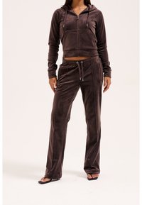 Femme portant un sweat à capuche zippé en velours marron foncé et un pantalon à cordon assorti avec des sandales noires à bout ouvert, debout sur fond blanc.