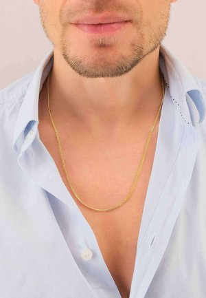 Mann mit leichtem Stoppelbart, der ein offenes hellblaues Hemd und eine dünne goldene Halskette um den Hals trägt.