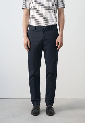 Person trägt schwarze Slim-Fit-Hose, schwarze Schnürschuhe und ein weißes kurzärmeliges Hemd mit dünnen schwarzen horizontalen Streifen.