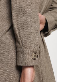 Brun herringbone-kappaärm med knappstängning, strukturerad manschett och ett mjukt, texturerat tyg med subtil lyster.