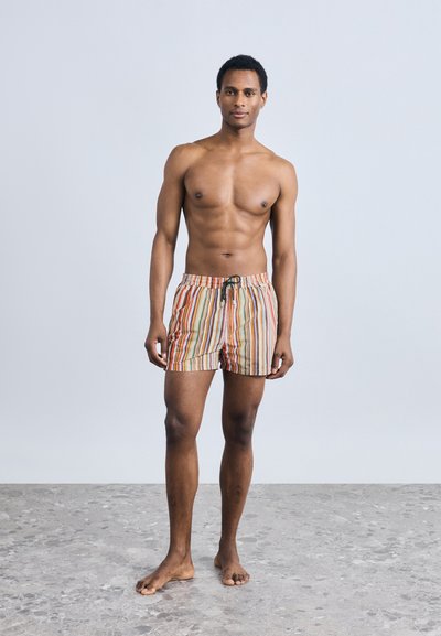 Ριγέ μαγιό σε πορτοκαλί, πράσινο και κίτρινο, με ελαστική μέση και μαύρο κορδόνι, που φοριέται από έναν άνδρα που στέκεται ξυπόλητος.