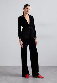 Black velvet blazer with a deep V-neck, paired with matching velvet trousers. Bold red shoes add contrast. Smooth texture and fitted design.