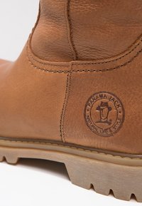 Gros plan sur une botte en cuir marron montrant des coutures détaillées et le logo Panama Jack en relief sur le côté, au-dessus d'une semelle beige robuste.