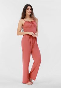 Mono de color rosa ribeteado, con tirantes finos, cuerpo fruncido y pantalones de pierna ancha con un dobladillo acampanado. Los pies están descalzos.