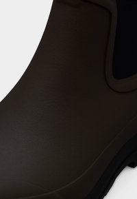 Bruine rubberen enkellaars met een gestructureerde afwerking, een zwart elastisch zijpaneel en een dikke zwarte zool voor verbeterde grip.