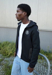 Sort zip-hoodie med et hvidt logo på brystet, med frontlommer med lynlås og en kontrasterende hvid inderside. Lyse denim shorts.
