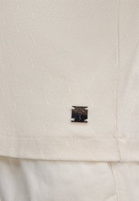 Cremfarbener Stoff mit Zickzackmuster, versehen mit einem kleinen silbernen Metallic-Emblem an der Seite, das mit vier Nieten befestigt ist.