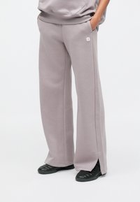 Χαλαρό φούτερ παντελόνι από απαλό, ανοιχτό γκρι ύφασμα με πλαϊνές σχισμές στο τελείωμα, με μικρό ροζ λογότυπο στο μπροστινό μέρος. Φορεμένο με μαύρα παπούτσια.