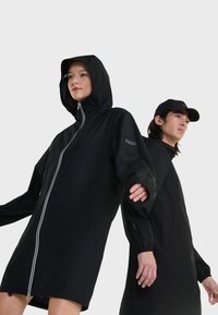 Giacca nera impermeabile con zip completa, cappuccio e polsini elasticizzati; presenta un logo discreto sul braccio e tasche su entrambi i lati.