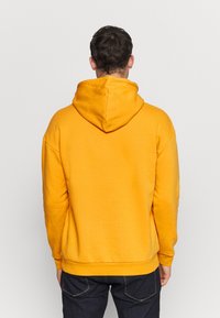 Gul hættetrøje med snøre i hætten, lange ærmer og ribstrikkede manchetter. Fremstillet af blødt stof, vist bagfra med en afslappet pasform.