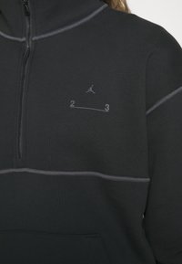 Sudadera negra con cremallera de cuarto, costuras grises, con un pequeño logo de Jumpman y la impresión "2 3" en el pecho. Textura de tela suave.