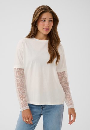 Jeune femme aux longs cheveux bruns portant un haut blanc avec des manches longues en dentelle visibles sous des manches courtes et un jean bleu clair.