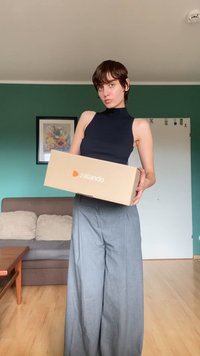 Zalando kartoninė dėžė natūralios rudos spalvos su oranžiniu logotipu. Modelis vilki prigludusį juodą be rankovių marškinėlį ir šviesiai pilkas platėjančias kelnes.