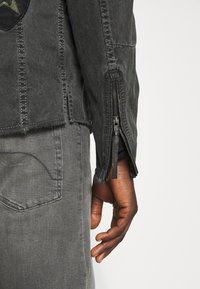 Musta, teksturoitu takki, jossa kontrastitikkaukset ja vetoketjulliset hihansuut, yhdistettynä harmaisiin farkkuihin, joissa näkyvä tasku.