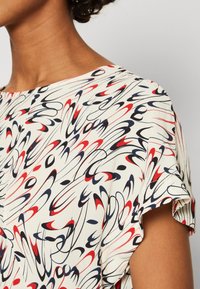Blusa de manga corta con escote redondo, con un fondo crema que presenta patrones abstractos en espiral en rojo, azul marino y negro. Textura suave.